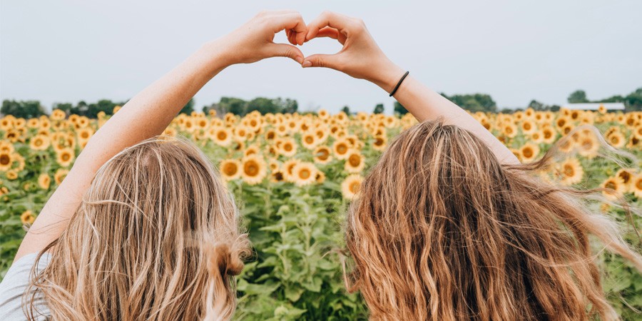 Two girls making a heart with their hands sitting in the field of sunflowers